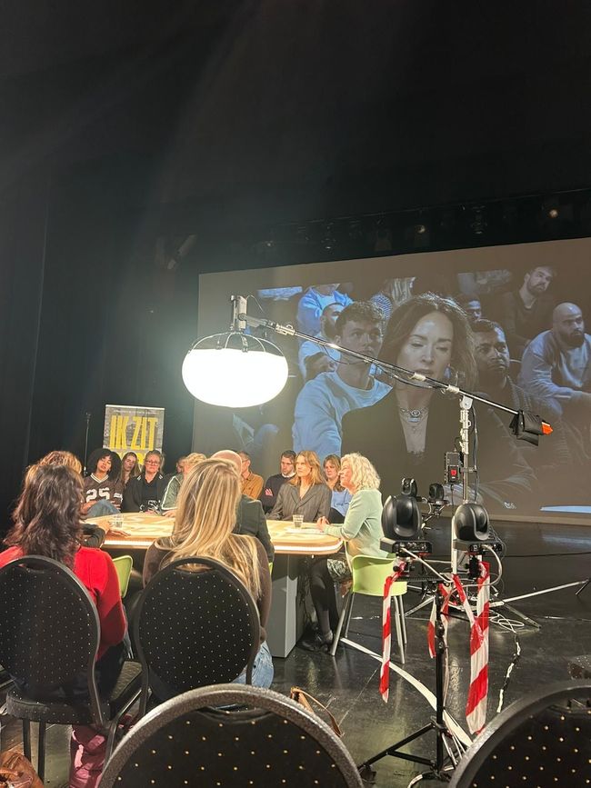 Rondetafelgesprek met panelleden aan een tafel, een camera-opstelling en een groot geprojecteerde spreker op een scherm.