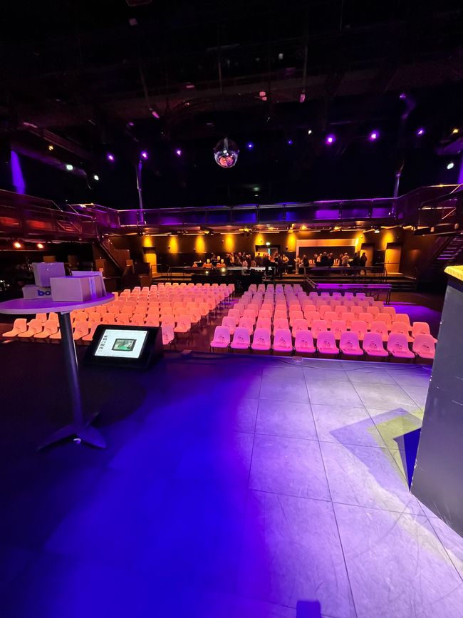 Een leeg theater met rijen roze stoelen onder paarse podiumverlichting, gezien vanaf het podium.