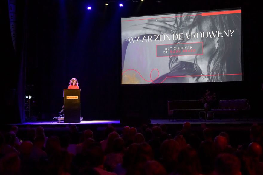 Spreker op een podium in een donker gebouw, met daarachter een groot presentatiescherm waarop een dia te zien is.