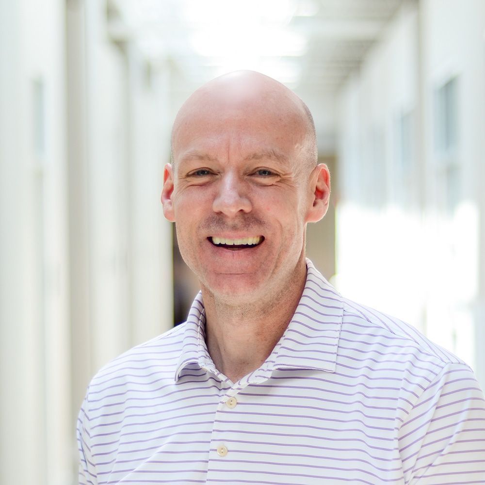 A black and white photo of a bald man smiling in front of a brick wall.