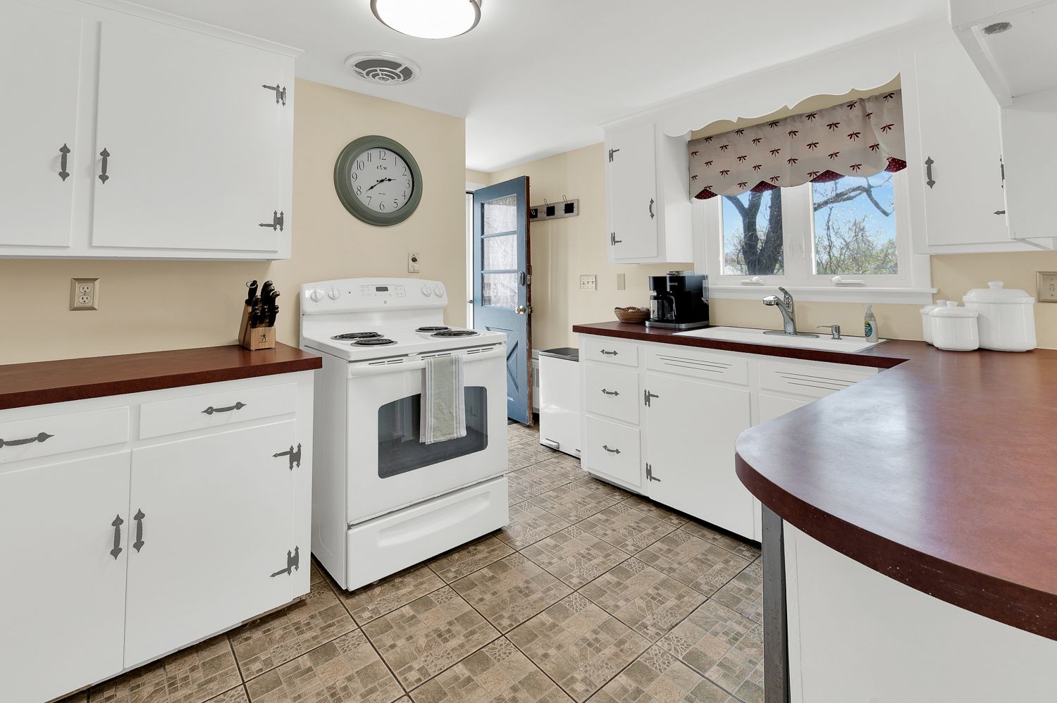 A kitchen with white cabinets , a stove , a sink and a clock on the wall.