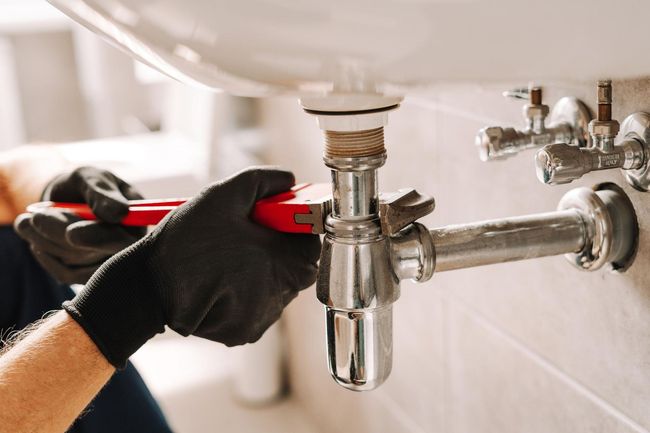 Gloved hands tightening a chrome sink drain pipe under a bathroom sink