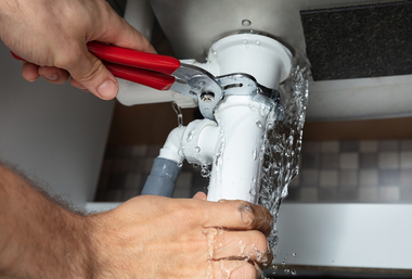 Plumber tightening a leaking pipe under a sink with red-handled pliers, water spraying out
