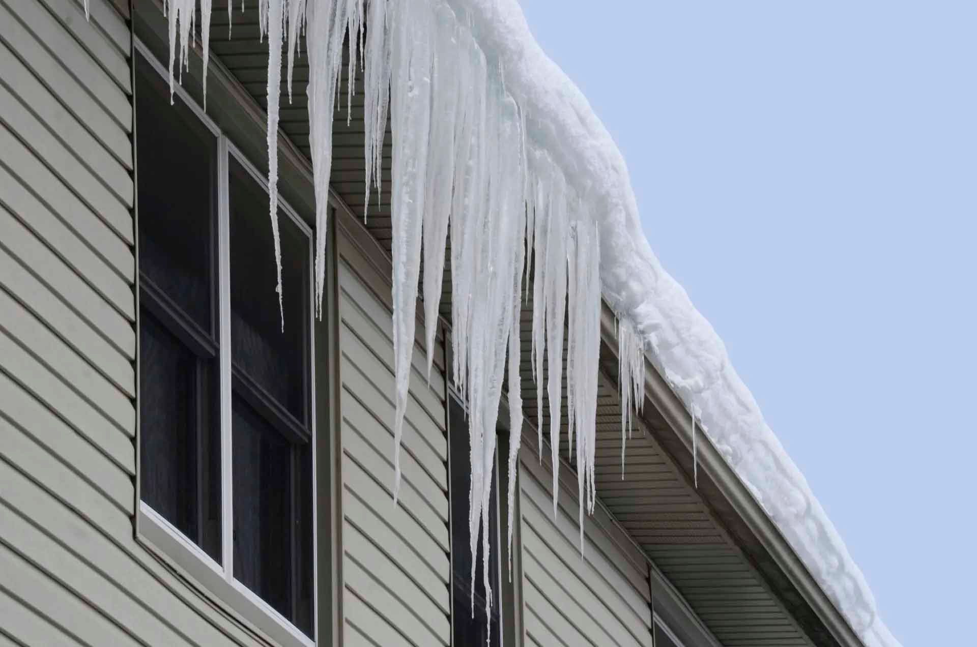 Des stalactites de glace pendent du toit d'une maison claire avec des fenêtres, sur un ciel bleu clair.