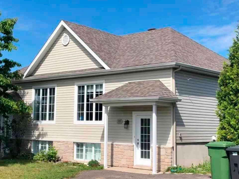 Maison à deux étages avec bardage beige, toit brun et fenêtres à cadre blanc. Entrée principale avec petit porche.