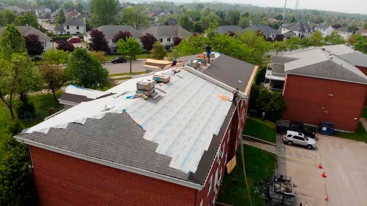 Des ouvriers posent le toit d'un immeuble en briques de deux étages, avec une vue partielle sur un quartier résidentiel.