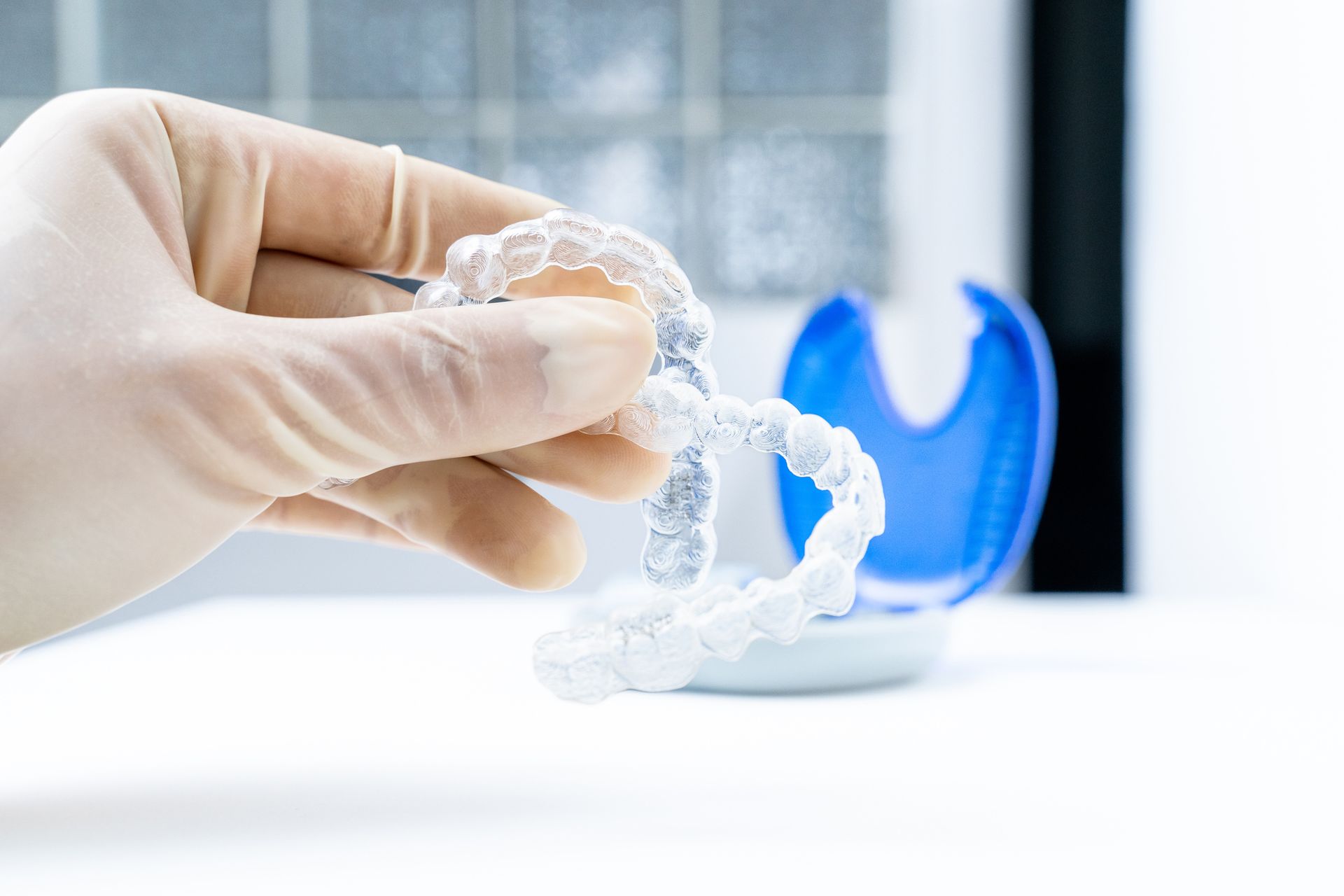 A gloved hand holding clear aligners, a blue case in the background, likely for dental work.