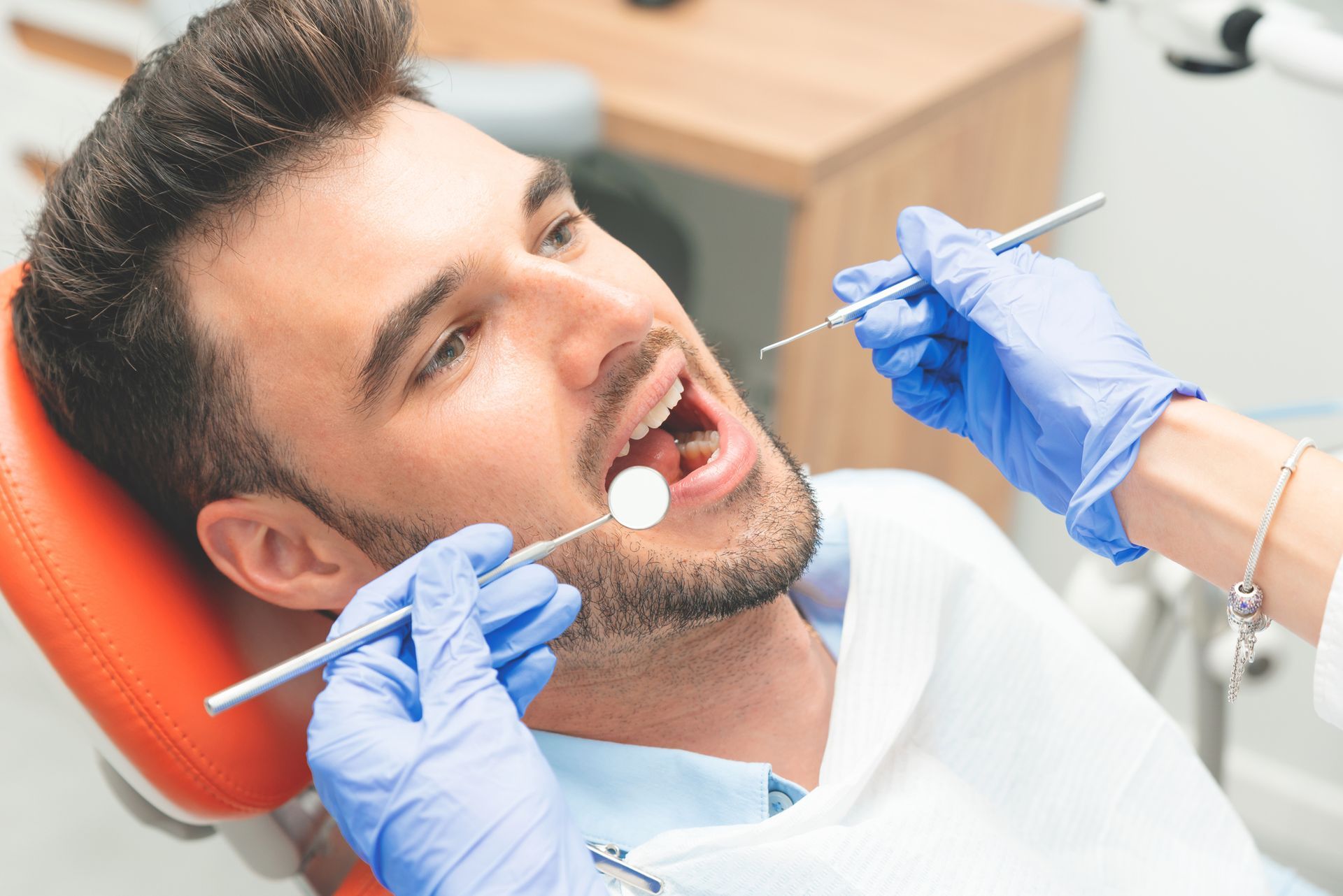 Man at dentist's office, mouth open for examination. Dentist in blue gloves uses tools to check teeth.