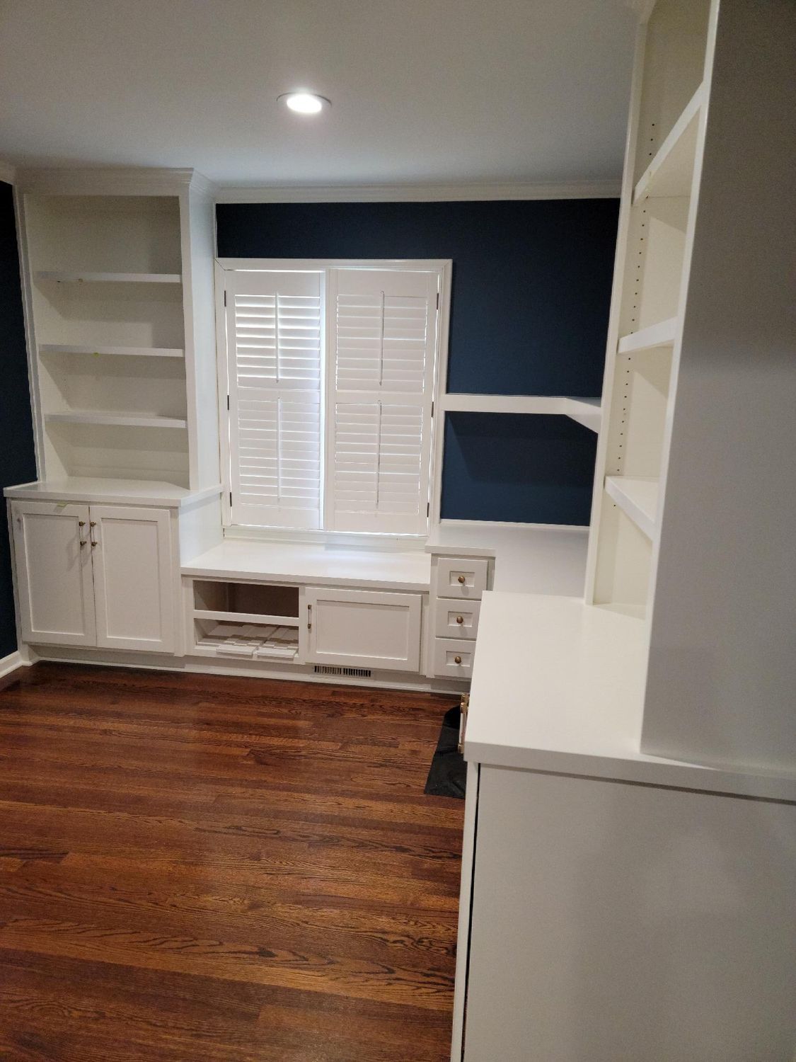 A room with hardwood floors , white cabinets , shelves and a window.