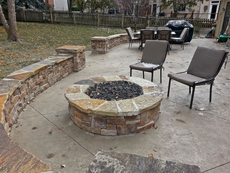 A fire pit is surrounded by chairs and a stone wall on a patio.