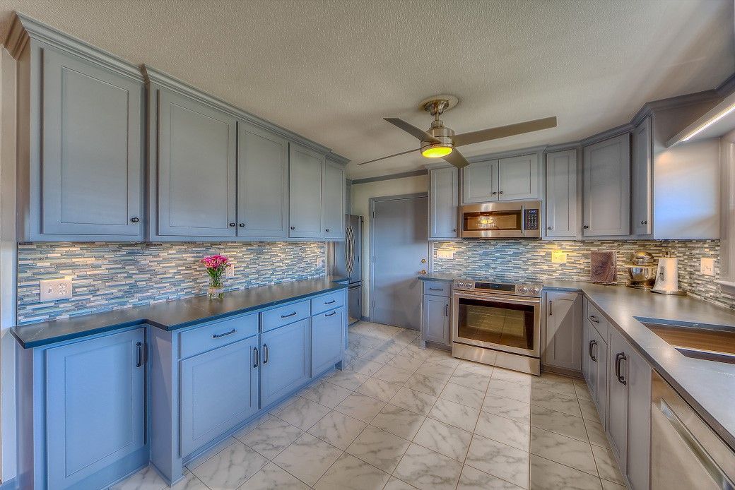 A kitchen with blue cabinets , granite counter tops , stainless steel appliances and a ceiling fan.