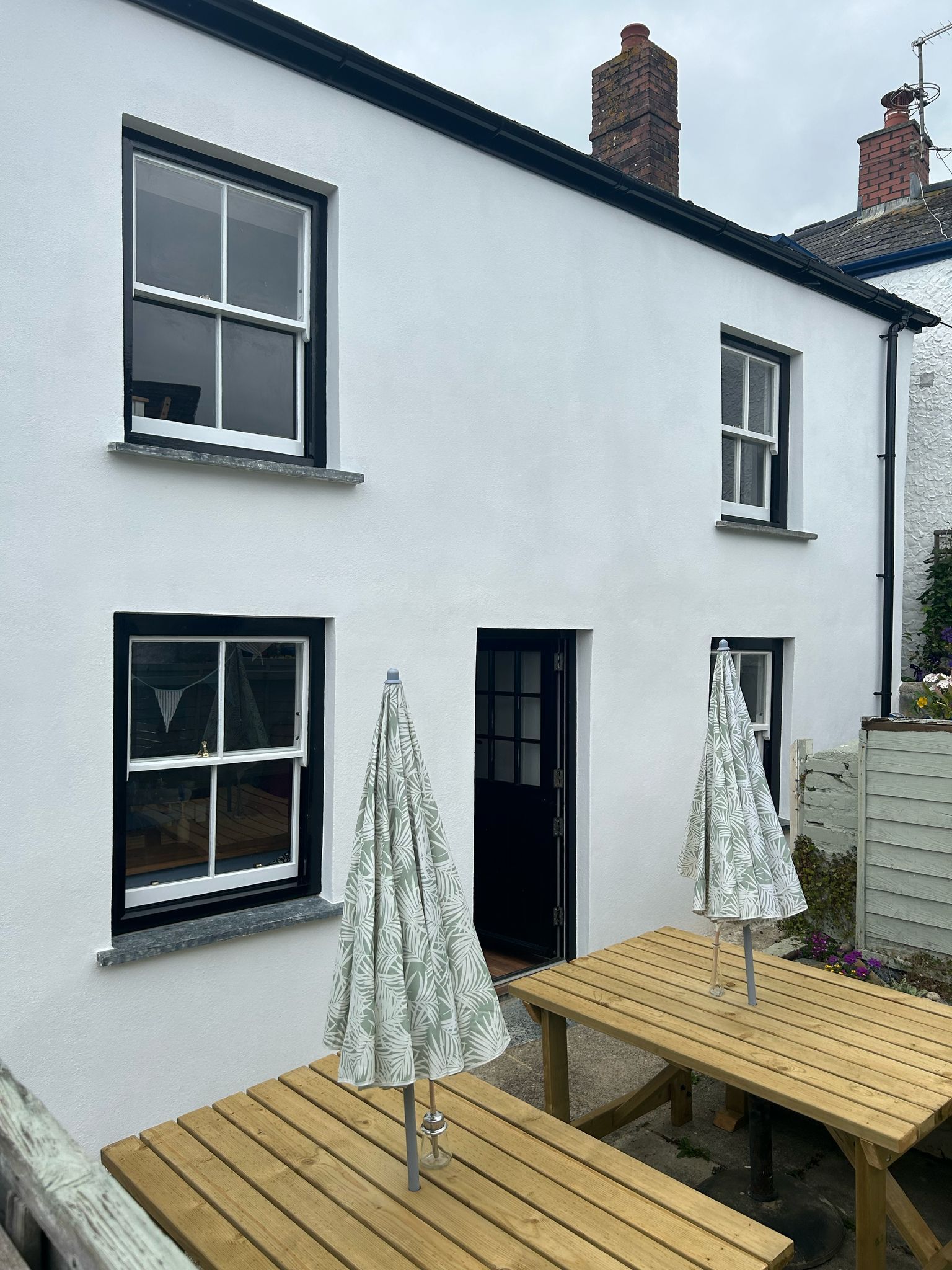 A white house with a wooden picnic table and umbrellas in front of it.