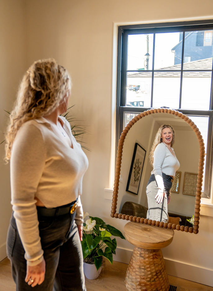 Woman looking in mirror; smiling, wearing a white sweater and black pants, standing near a window in a room.