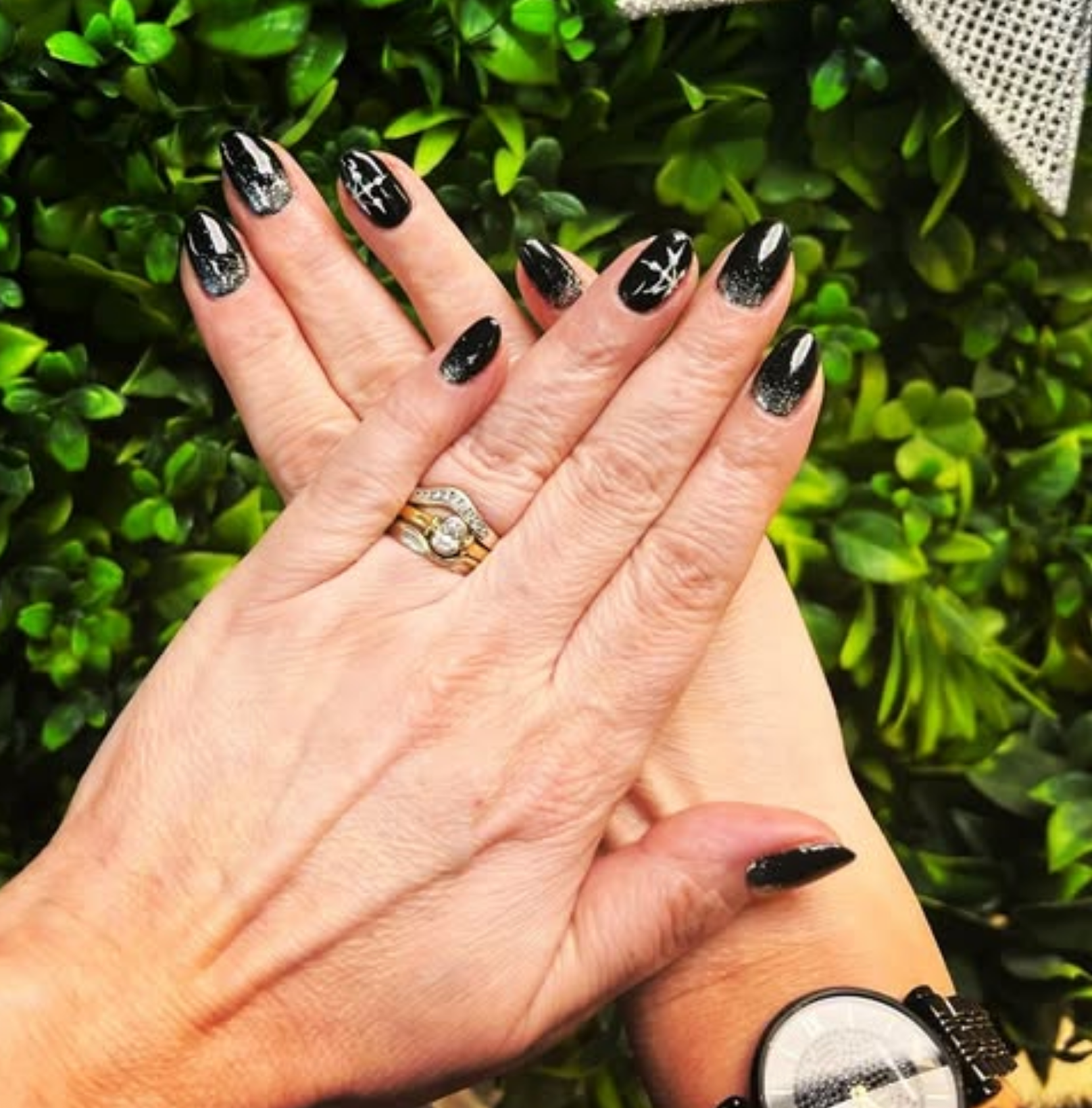 Hands with black and silver ombre nails, wearing rings, crossed in front of greenery.