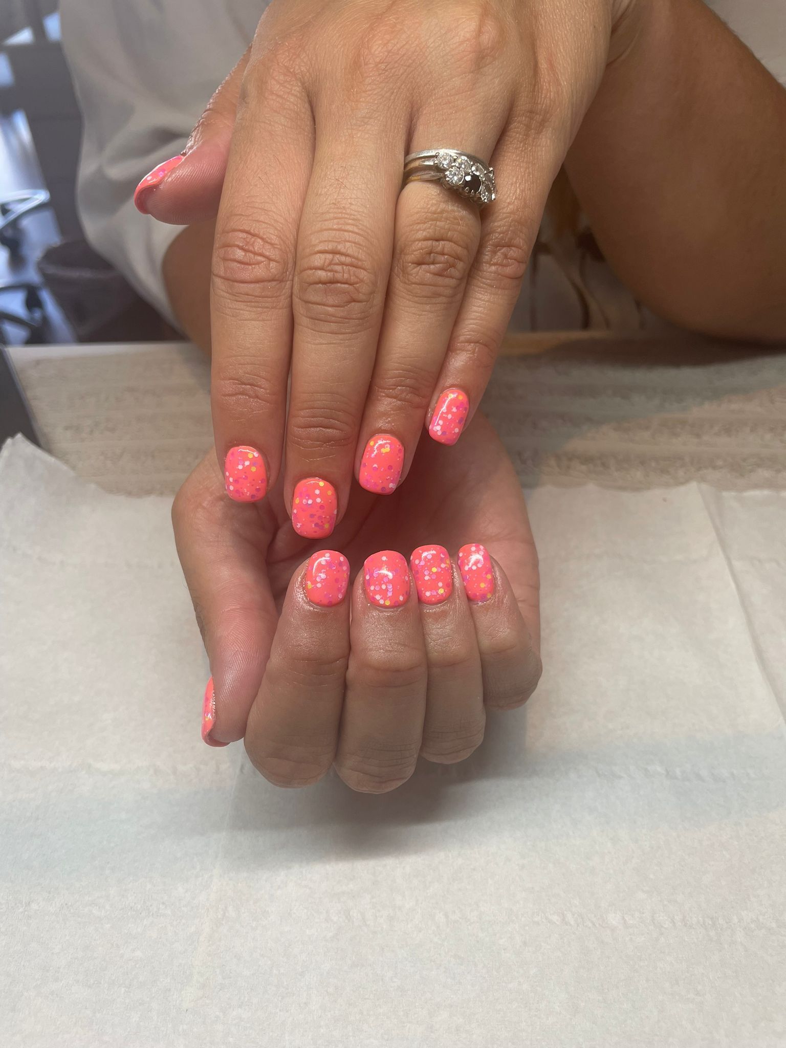 Hands with short, pink manicured nails, displayed on a white surface. One hand has a ring.