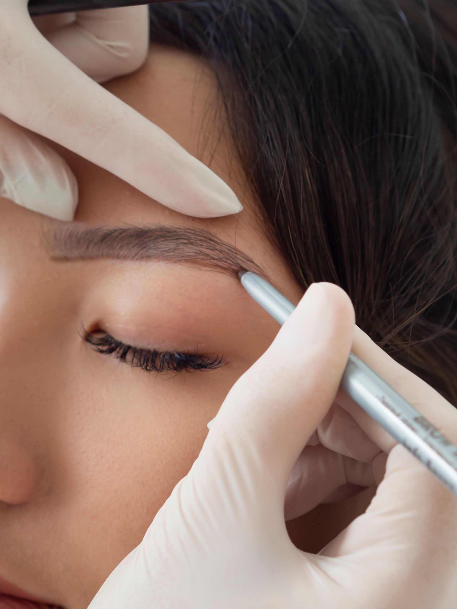 Eyebrow microblading procedure: gloved hand using a tool to draw a brow on a person's face.