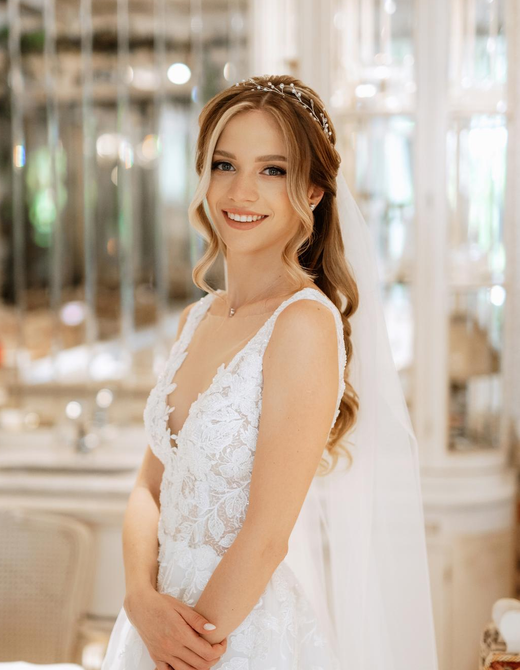 Woman in a white wedding dress smiles, with a floral headband, in a bright room.