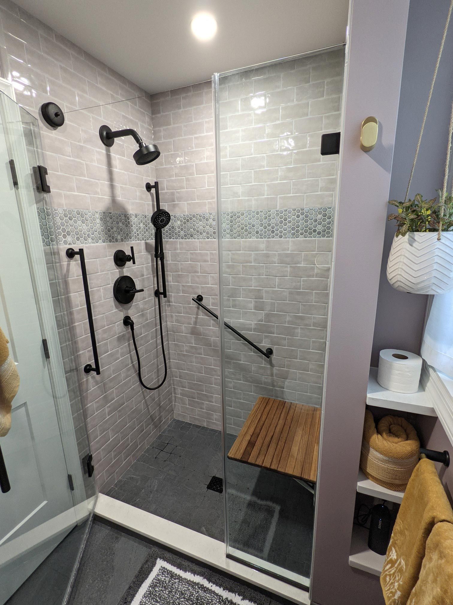 Shower with black fixtures, gray tile, glass door, wooden seat, and grab bar.
