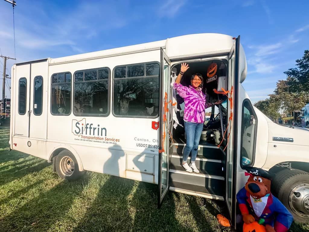 Person waves from a Siffrin bus doorway in an outdoor setting. The bus is white; the person wears a tie-dye shirt.