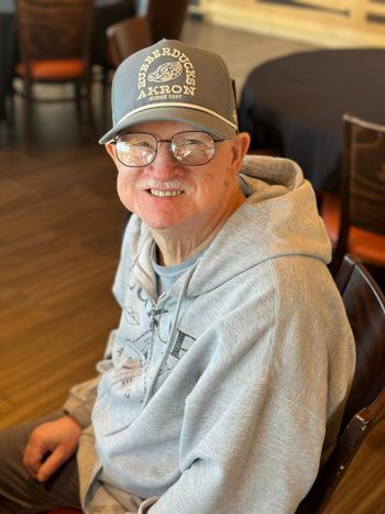 A smiling man in a gray hoodie and hat sits indoors. He wears glasses and has a friendly expression.
