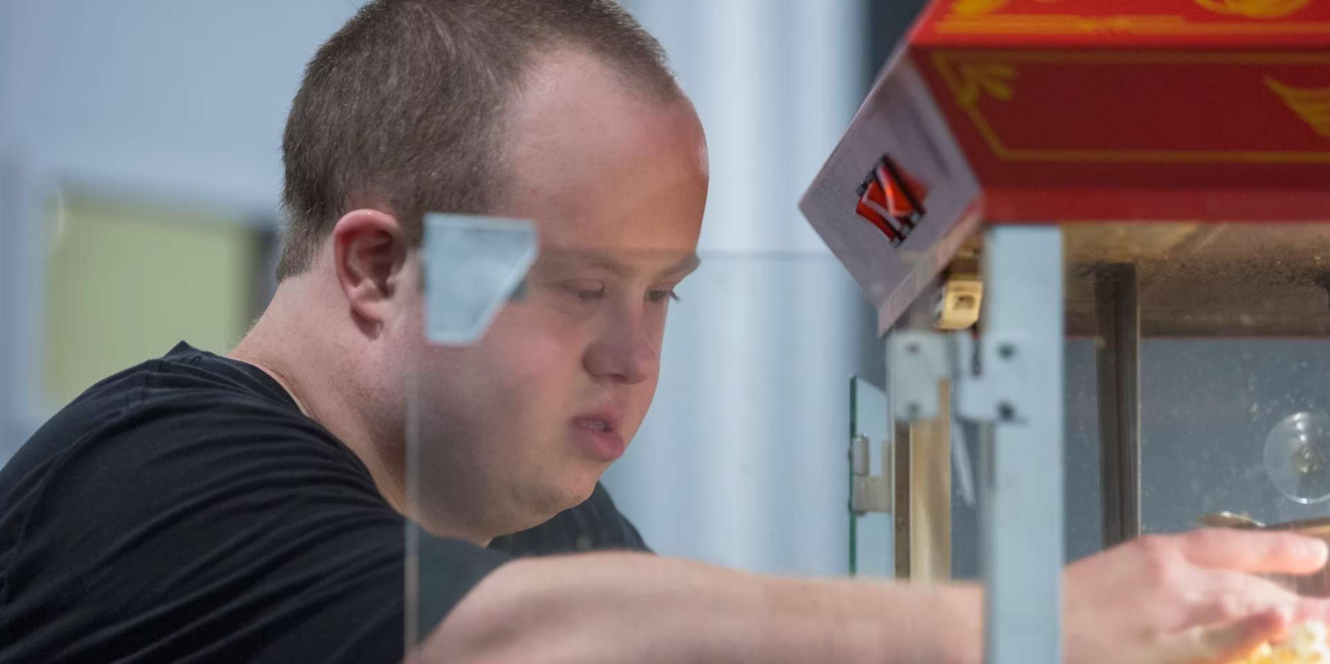 A man with Down syndrome tending a popcorn machine. He wears a black shirt and has a thoughtful expression.