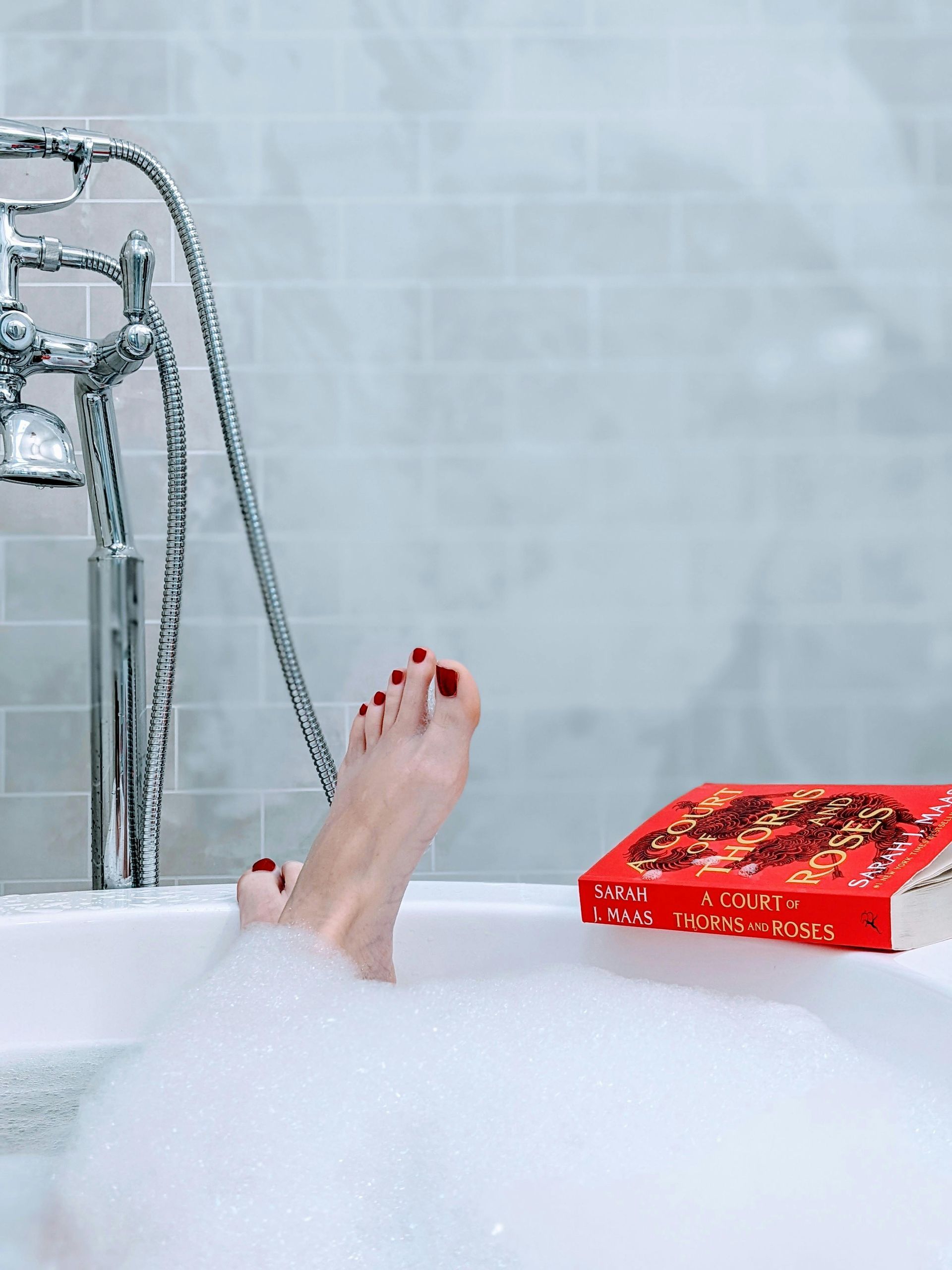 Feet with red toenails in a bubbly bathtub, a book resting nearby.