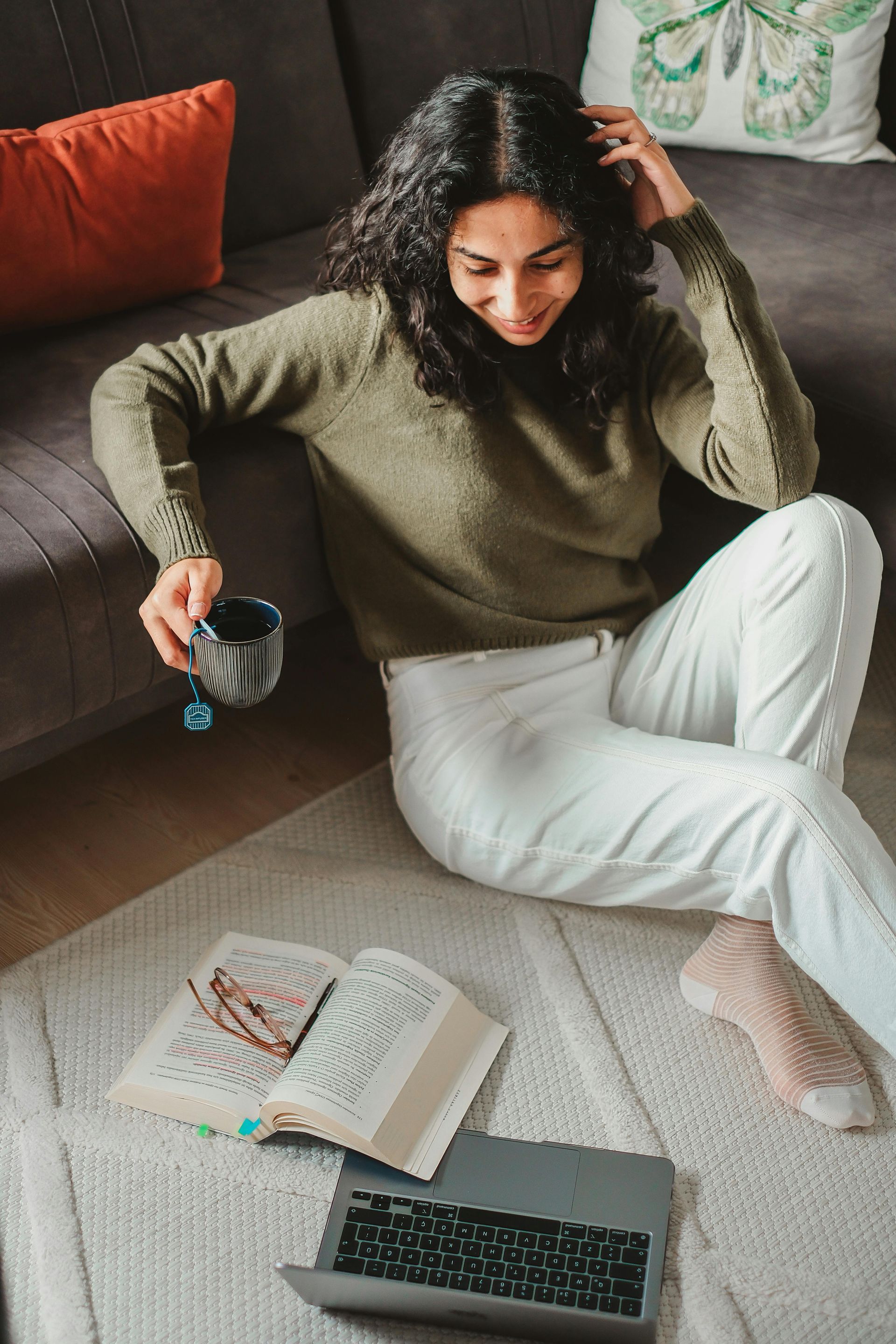 Woman seated on floor with book, laptop, and coffee cup, near a sofa.