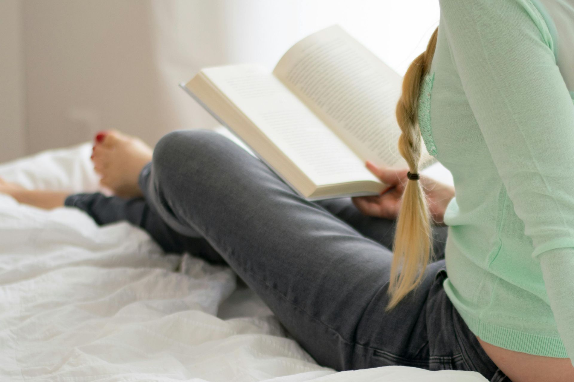Person reading a book while reclining on a bed, wearing a light green shirt and gray jeans, with a blonde braid.