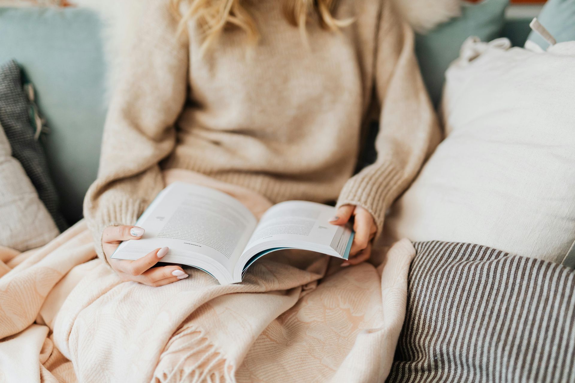 Woman reading a book on a couch, surrounded by pillows and a blanket.