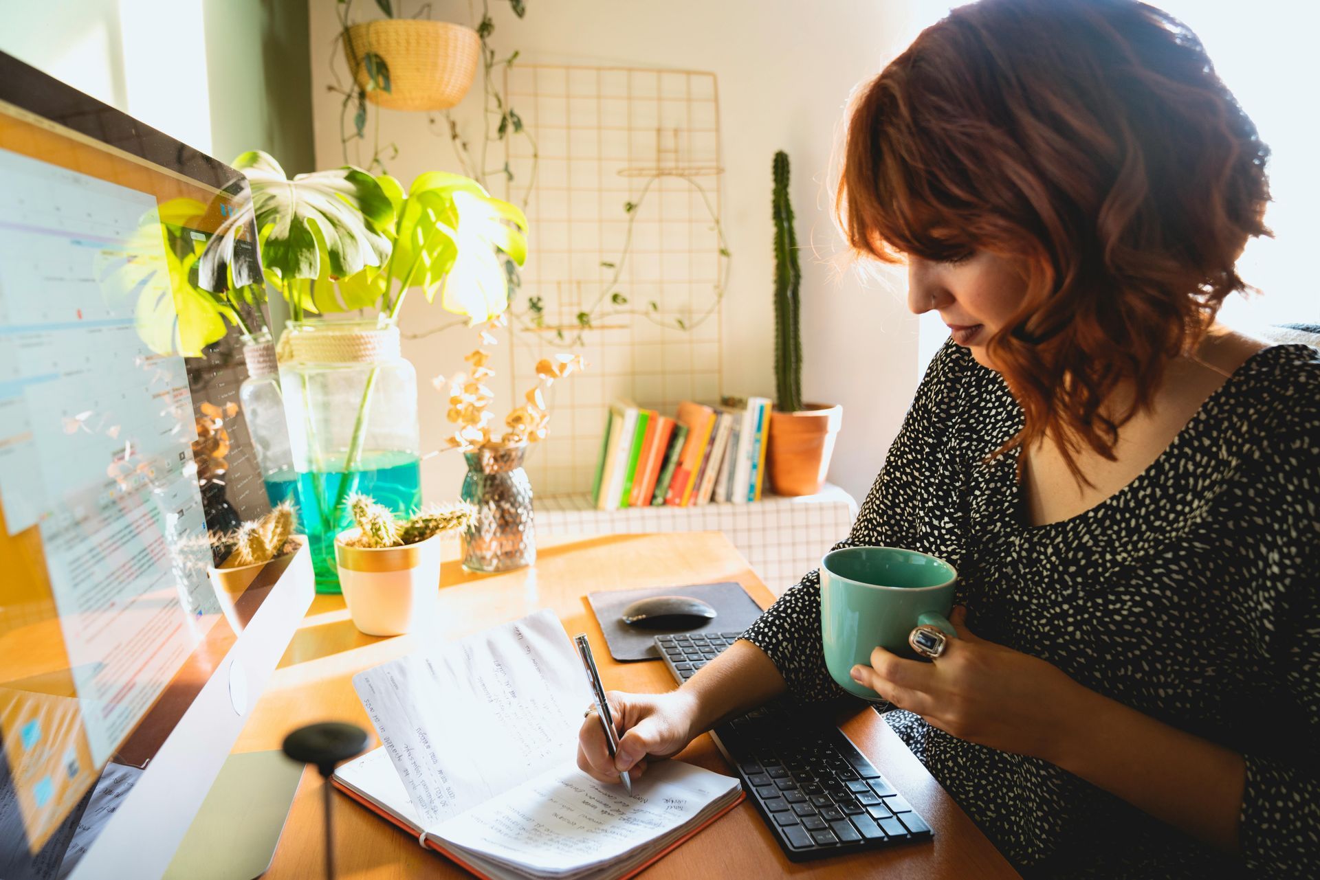 Woman writing at a desk, holding a mug, with a computer and plants nearby.
