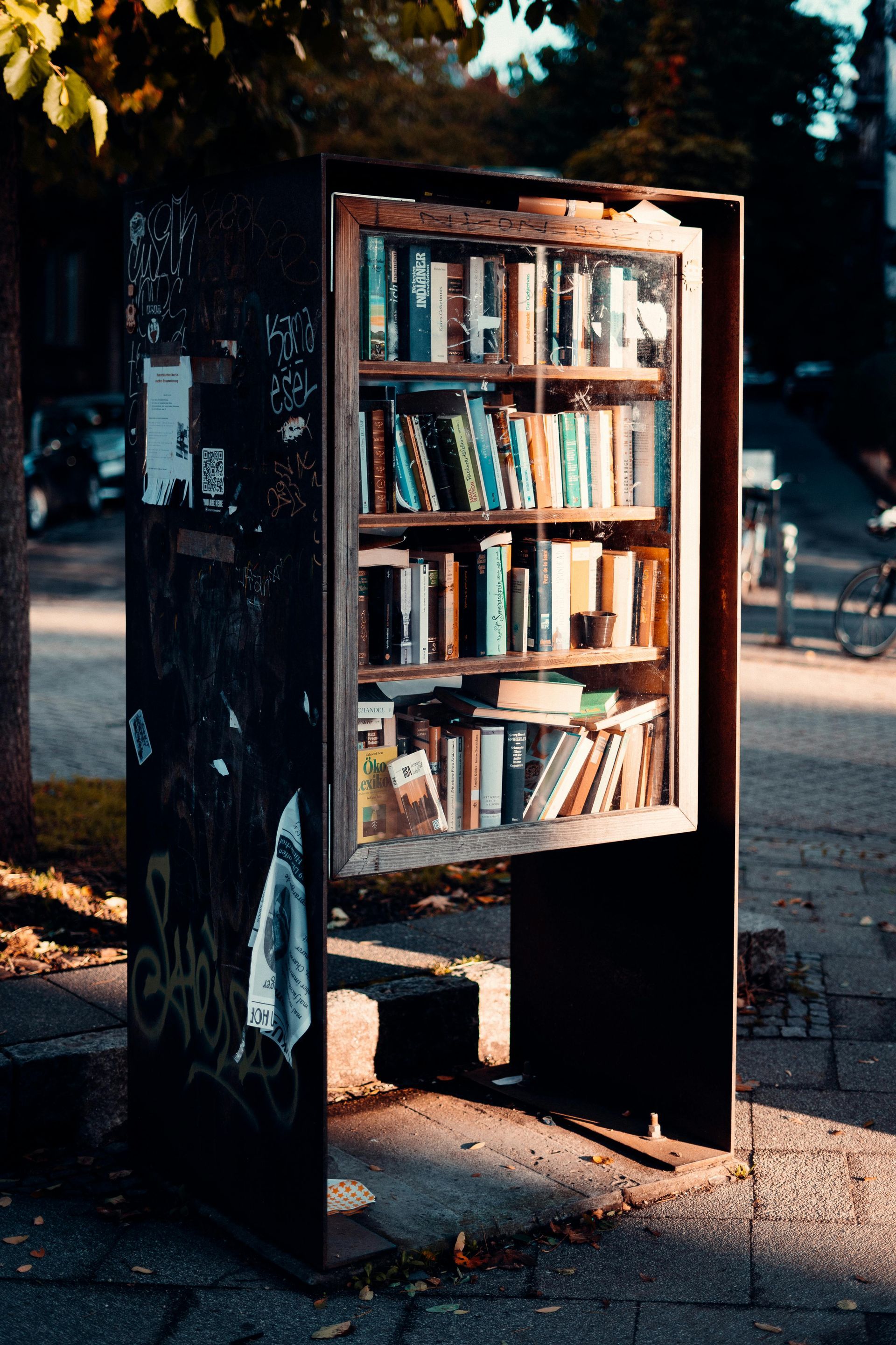 Book-filled outdoor library with glass door, on a sidewalk, in a shadowed, urban setting.