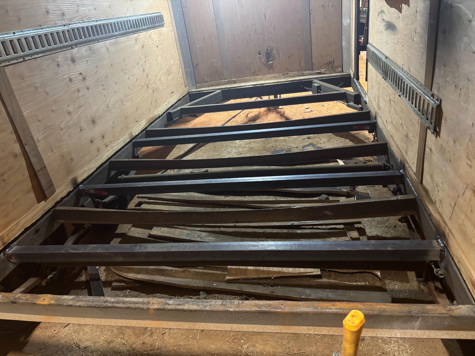 A metal trailer frame sits inside a wooden storage container, with miscellaneous metal pieces stacked below the frame.