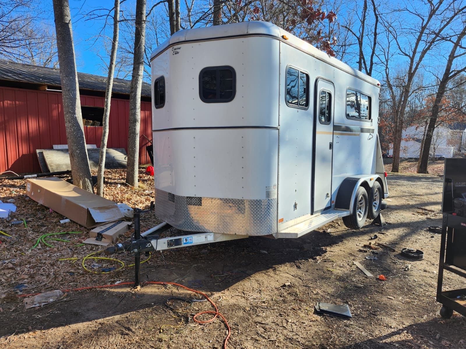 Silver horse trailer parked on dirt, with trees and a red building in the background.