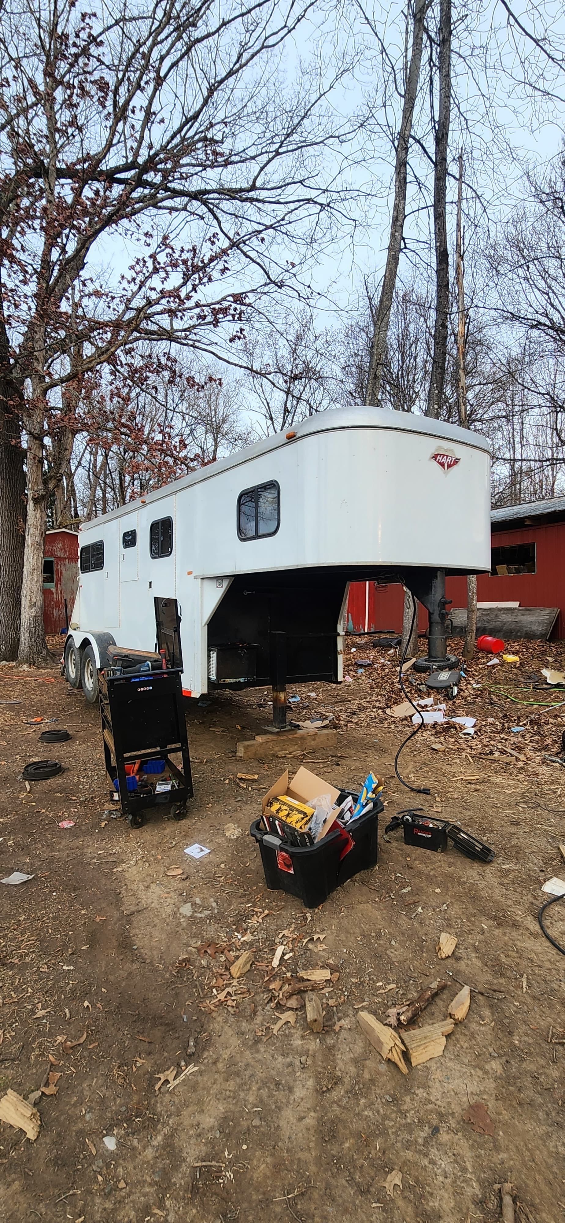 A white horse trailer parked in a yard with two containers in front. Trees and a red building are in the background.