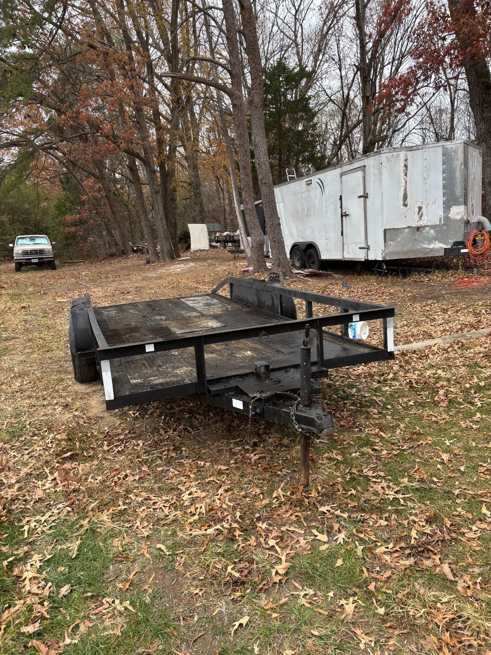 Black utility trailer in a grassy area with a white trailer in the background. Autumn leaves are scattered around.