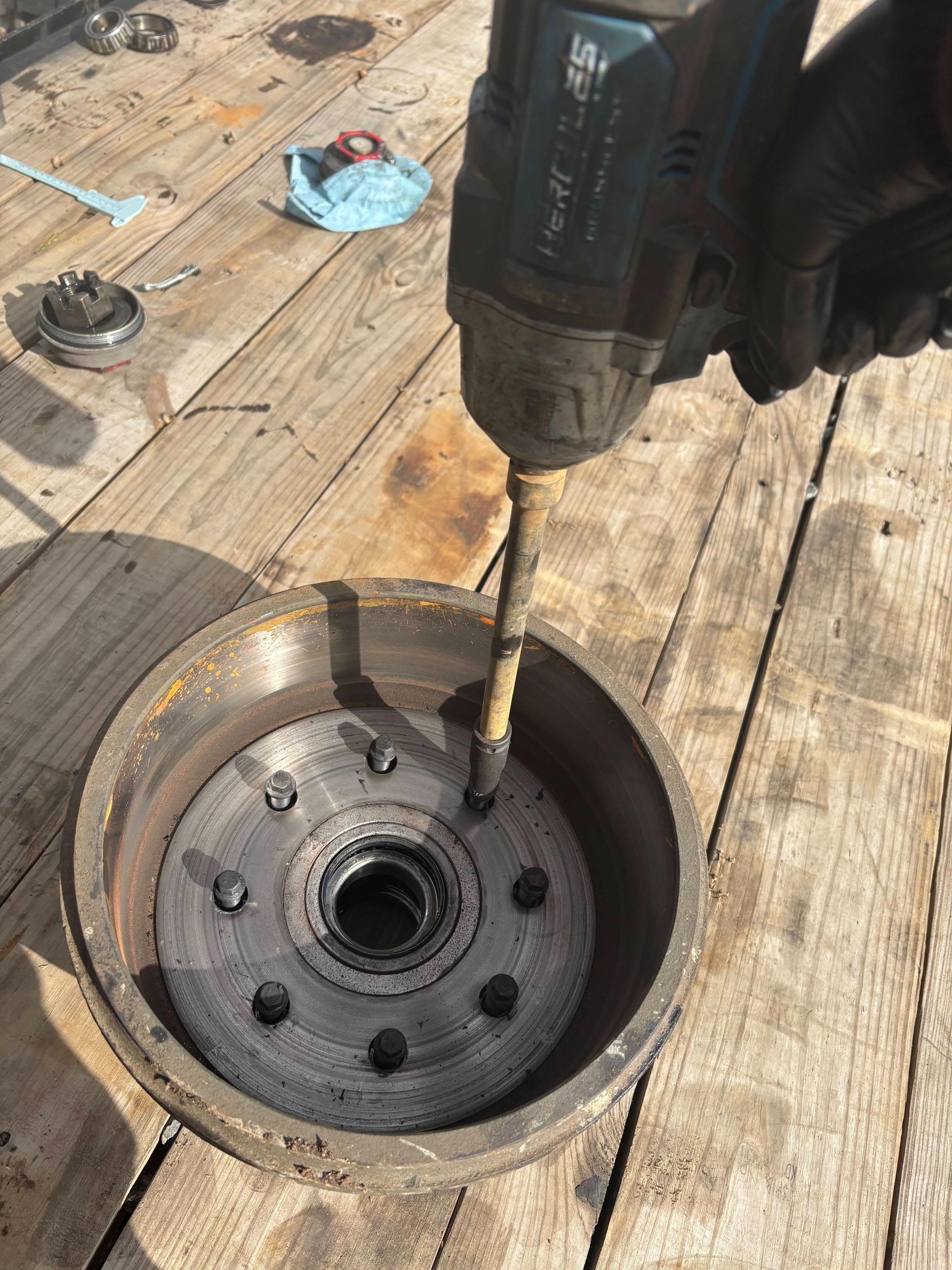 A gloved hand uses an impact driver to work on the hub of a metal brake drum resting on wooden planks.