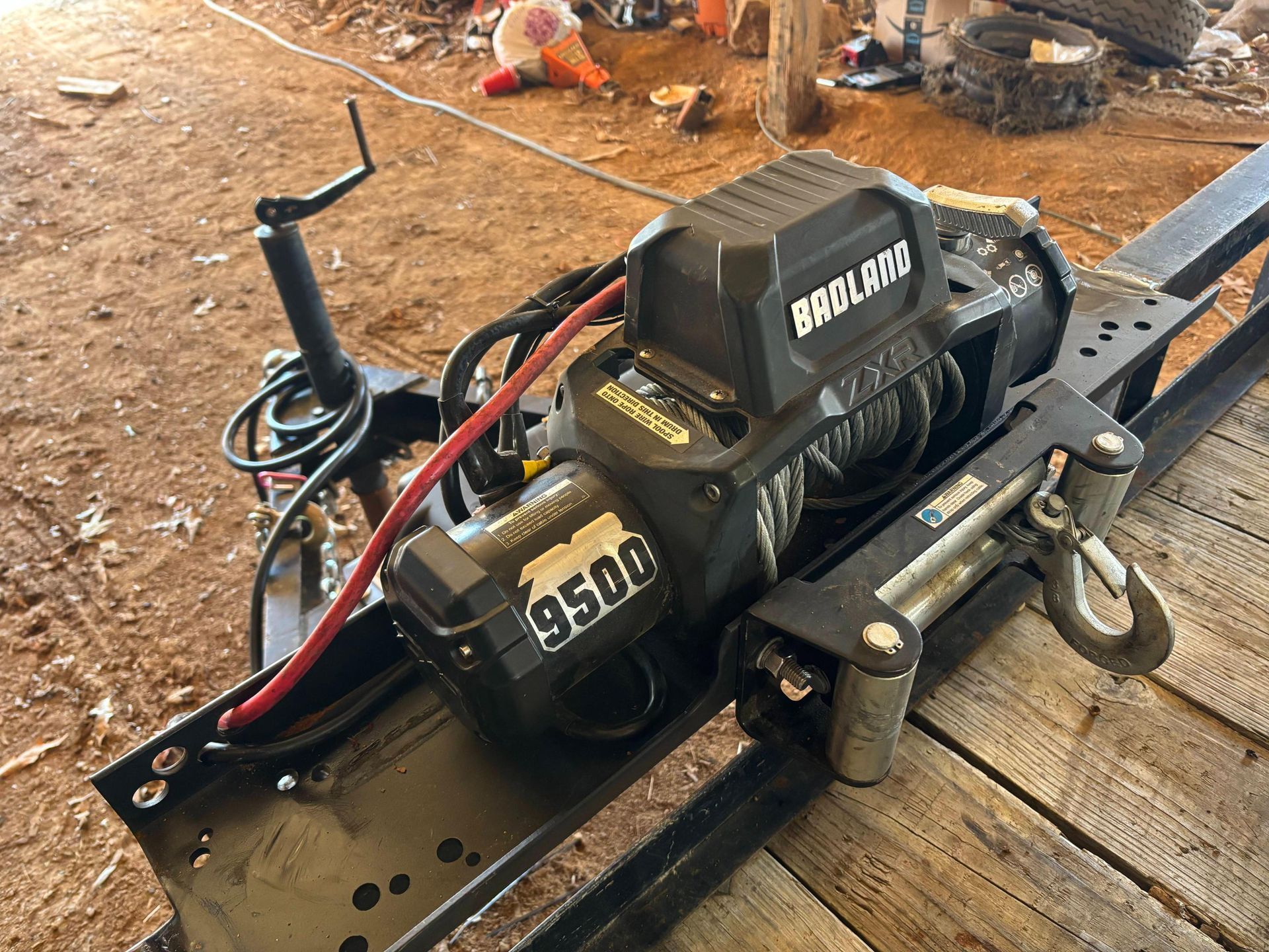 A black Badland 9500 winch mounted on a metal frame, sitting on a wooden surface in a dusty outdoor area.