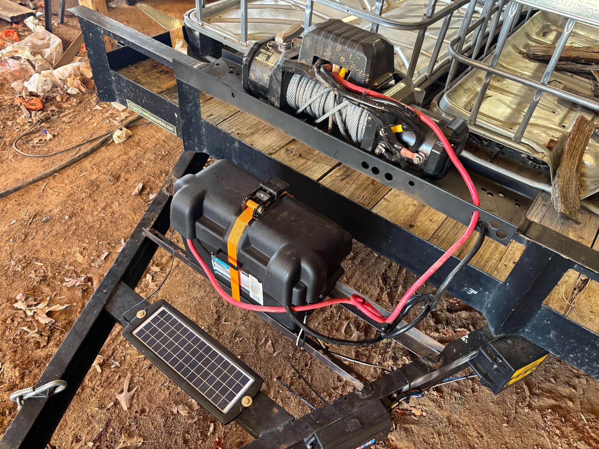 An electric winch, battery in a black case, and small solar panel mounted on the tongue of a metal utility trailer.