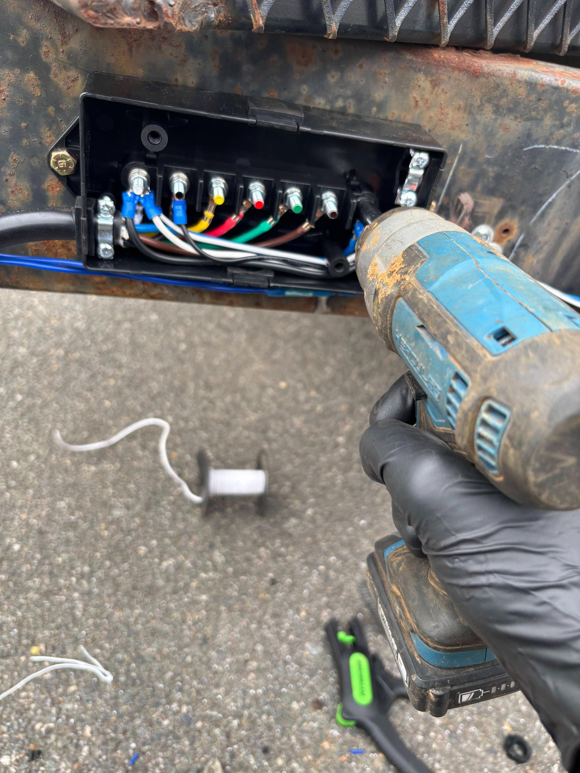 A gloved hand holds a drill over a trailer junction box with multicolored wires being connected to a terminal block.