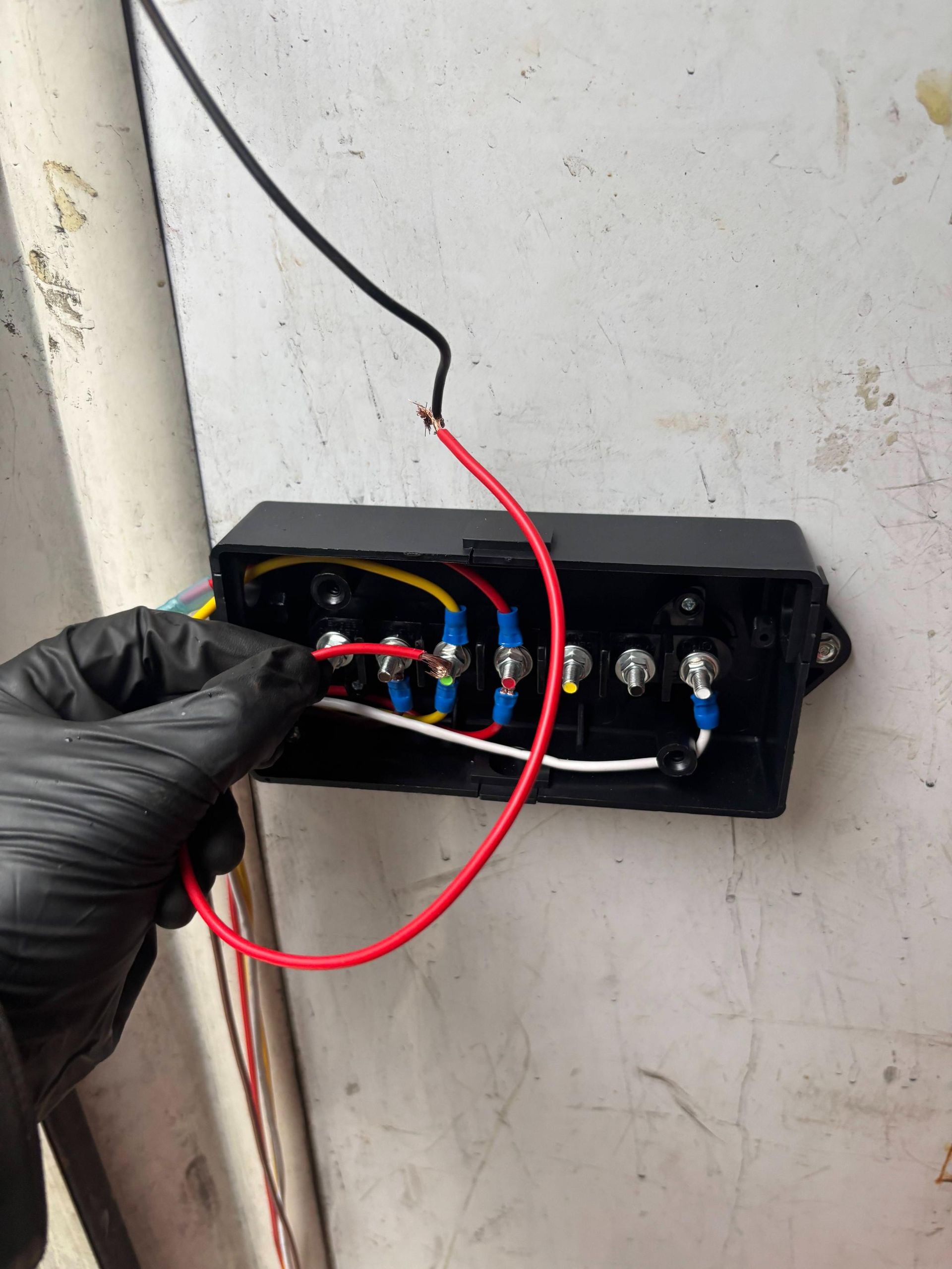 A gloved hand holds a red wire near an open electrical junction box mounted on a wall with various colored wires attached.