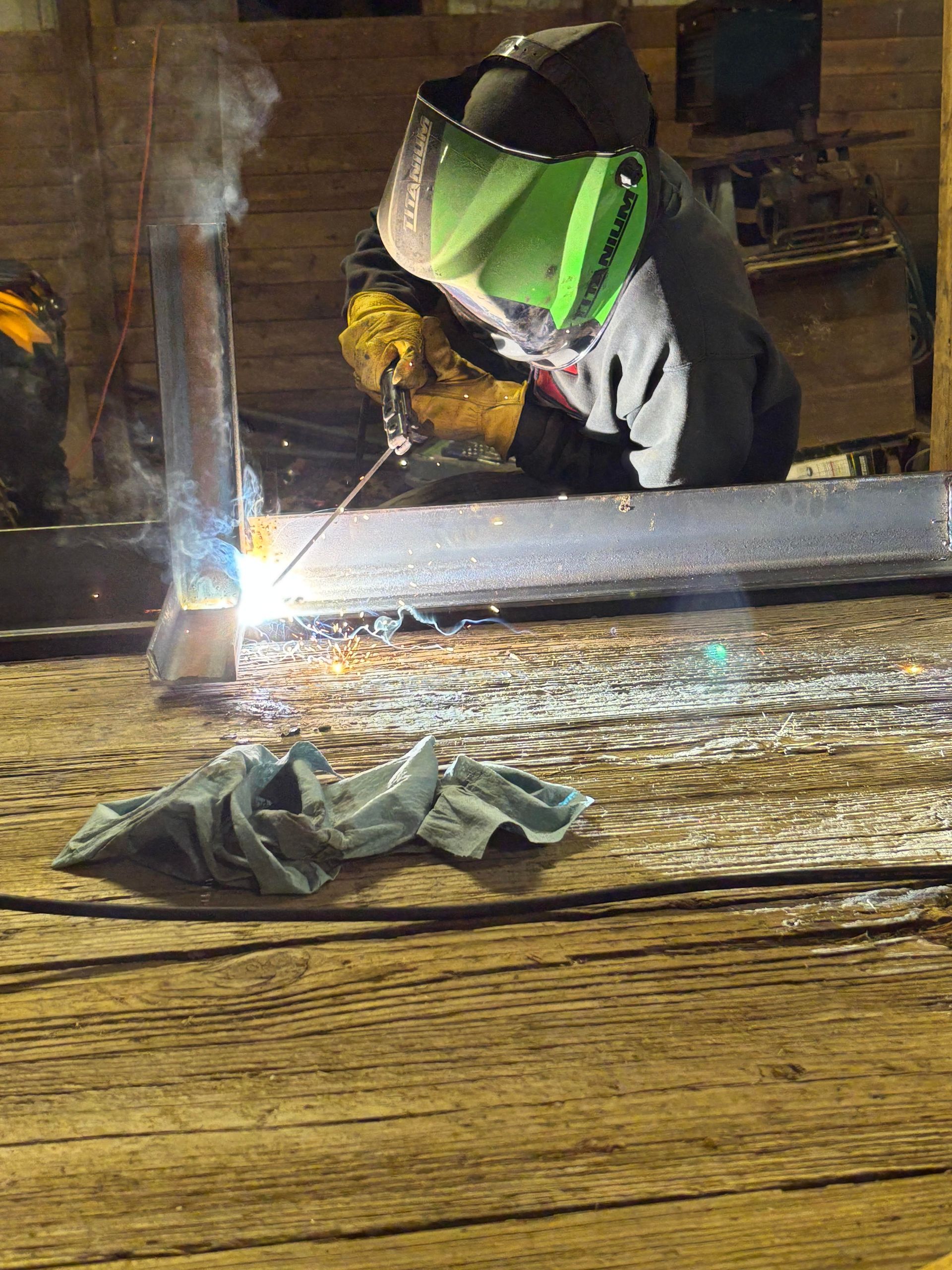 Welder wearing a green-tinted helmet welding metal in a dim setting; smoke and sparks visible.
