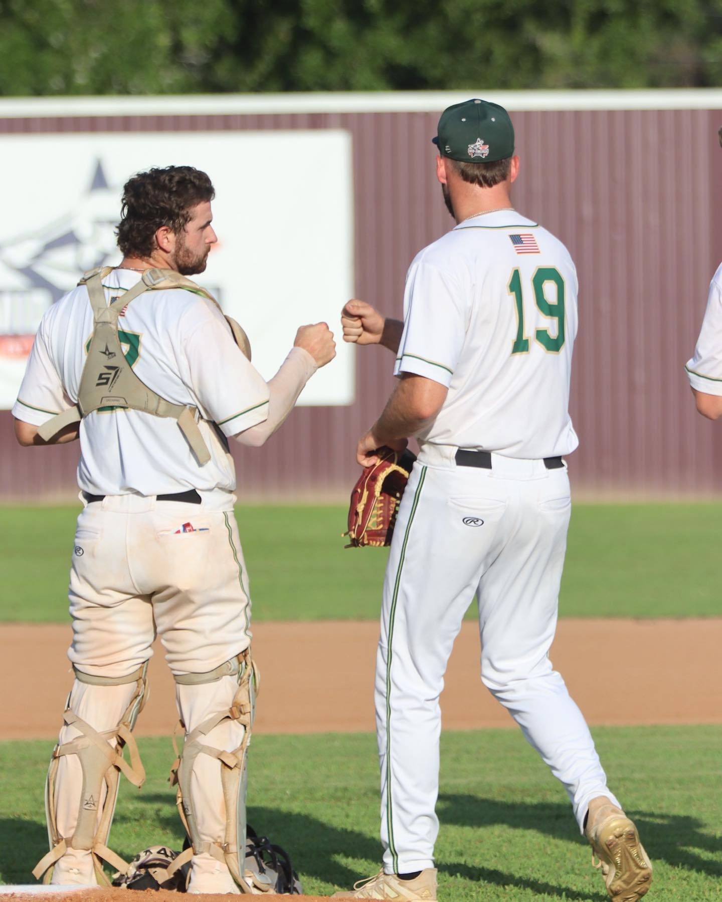 Two baseball players in white and green uniforms bump fists on a baseball field.