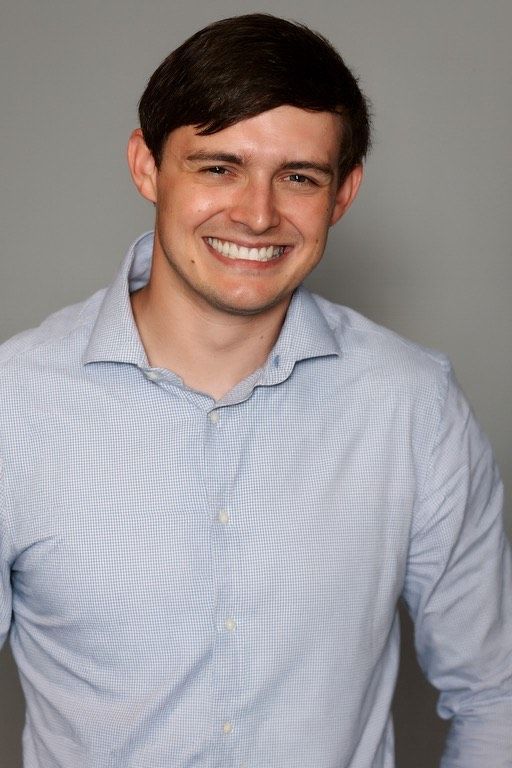 Man smiling, wearing a light blue patterned button-down shirt, against a gray background.