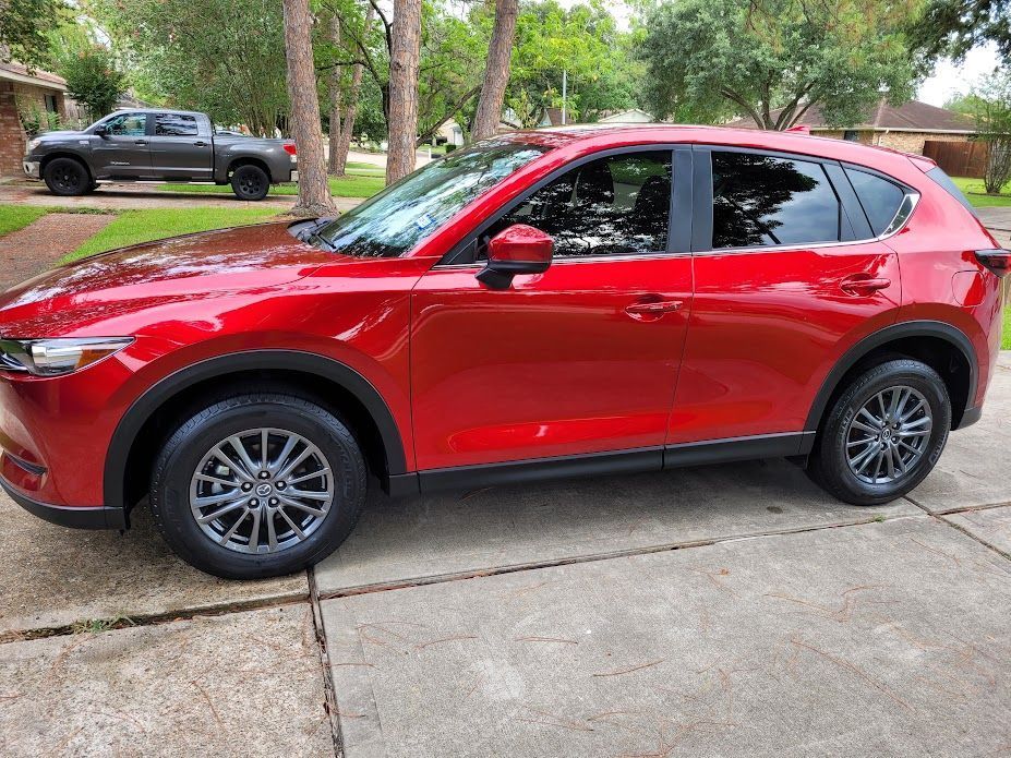 Red Mazda CX-5 SUV parked on a concrete driveway in front of a house, with a gray truck in the background.