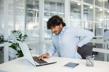 A man is sitting at a desk using a laptop computer while holding his back in pain.
