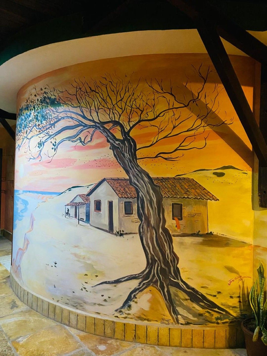 Mural representando uma árvore castigada pelo vento e duas casas em uma praia arenosa, pintado em uma parede curva com cores quentes.