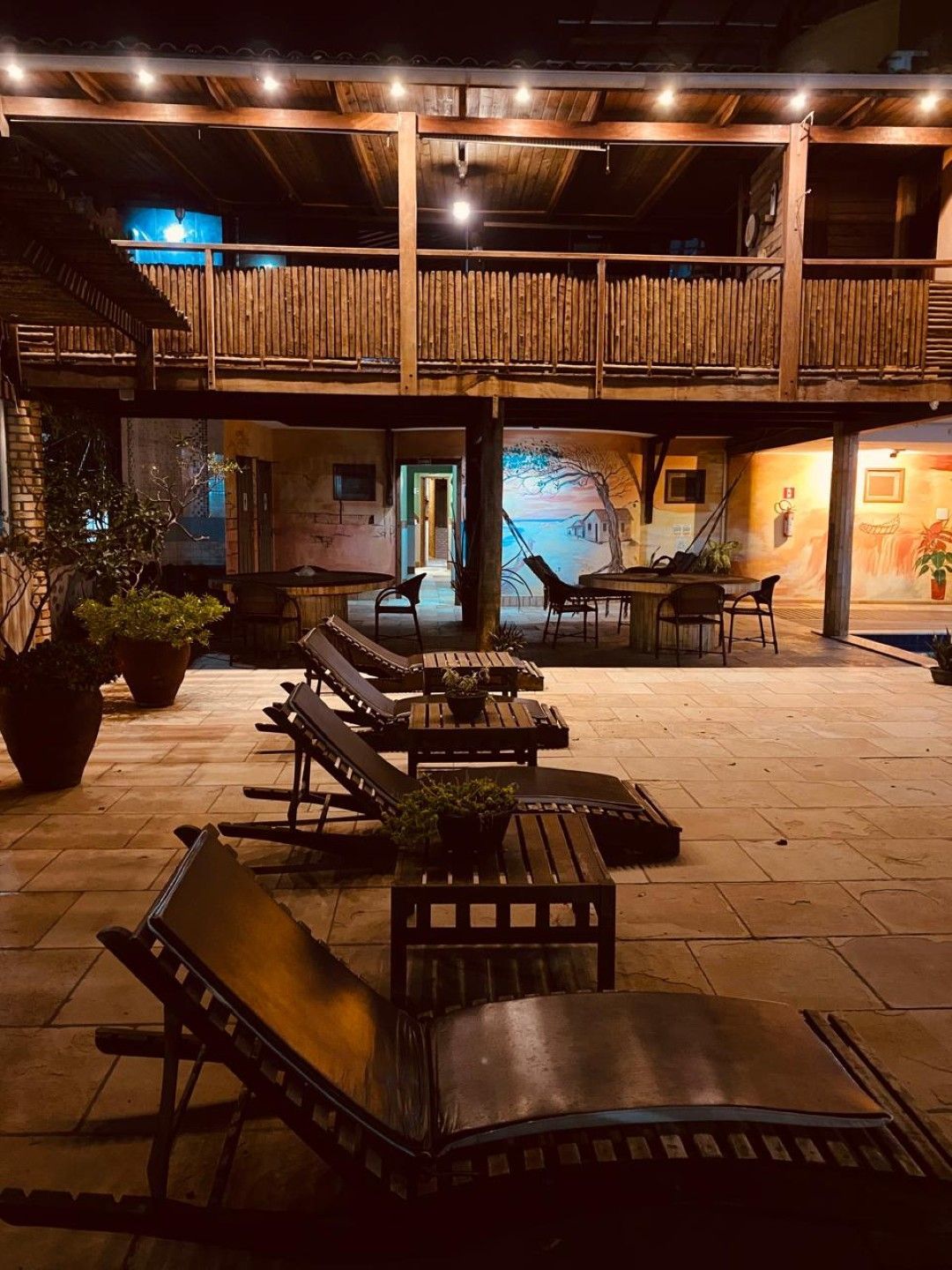 Área externa com espreguiçadeiras de madeira, mesas e varanda suspensa iluminada à noite.