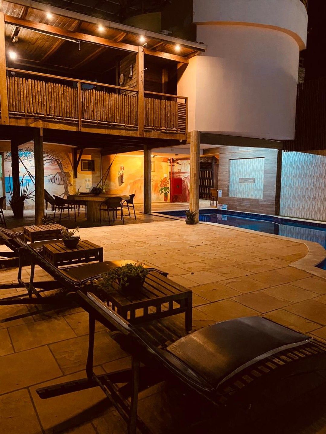 Pátio com espreguiçadeiras, piscina e varanda de madeira à noite.