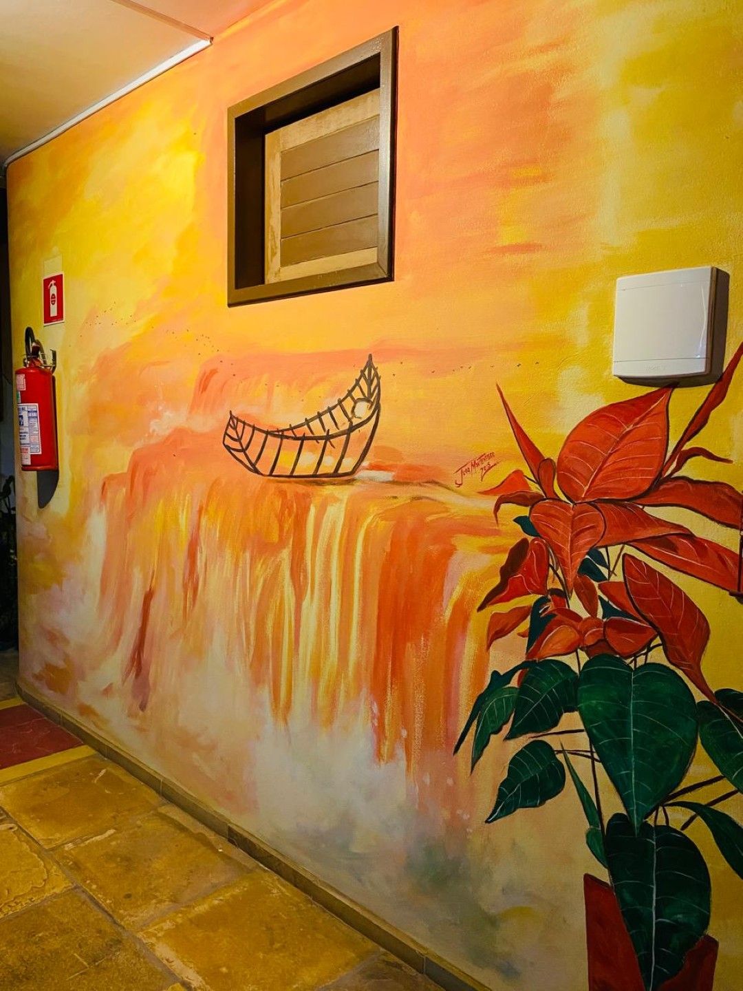 Corredor com mural colorido de um barco perto de uma cachoeira, extintor de incêndio e planta de poinsétia.