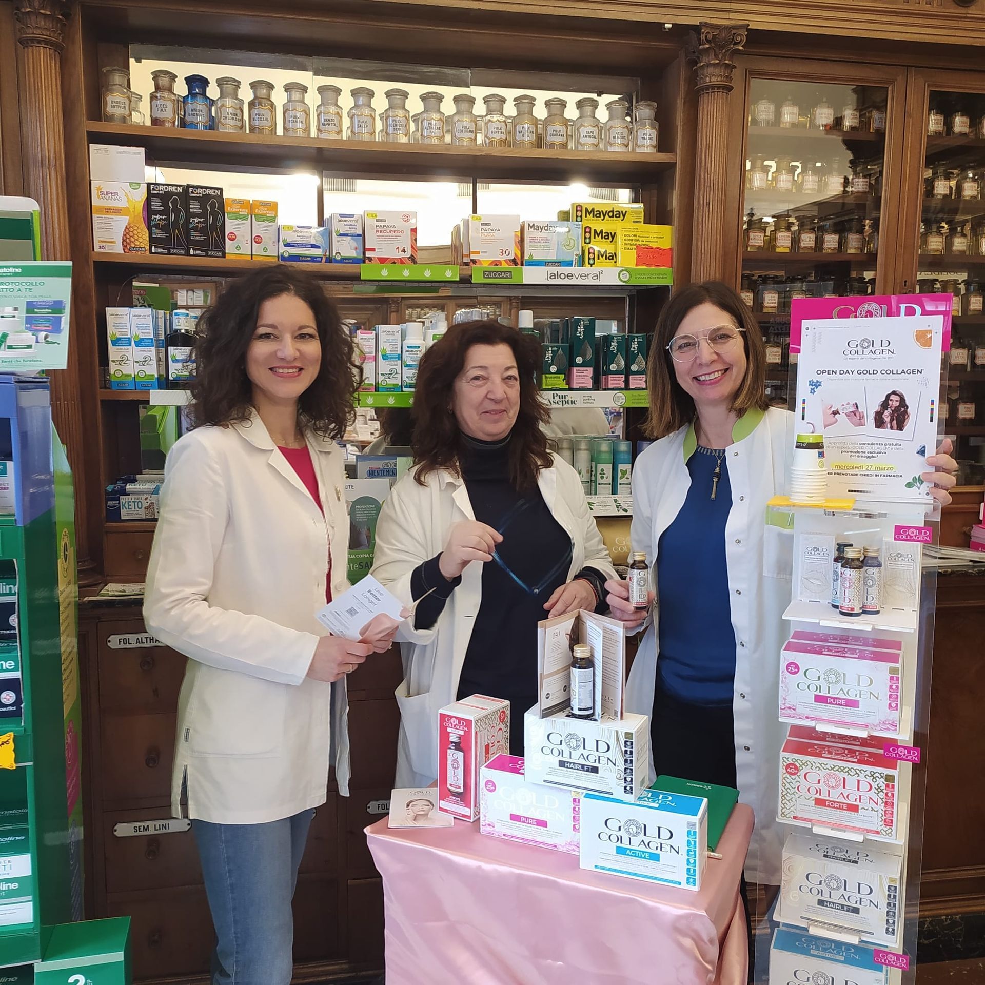 Tre donne in camice bianco sono in piedi in una farmacia, espongono i loro prodotti. Dietro di loro ci sono scaffali pieni di merce.